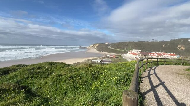 Ocean waves crash onto the sandy shore of Praia de Paredes da Vitria, with rocky cliffs in the background. The shimmering water and coastal breeze. Green grass and yellow flowers frame the view.
