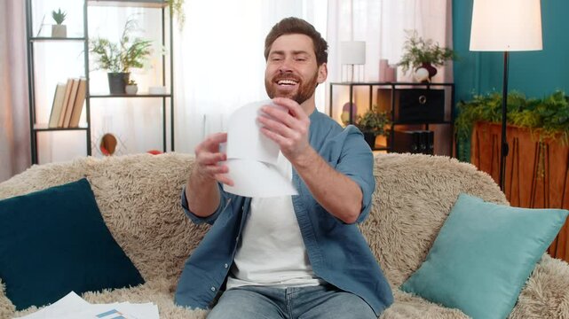 Young man at home sits on the sofa, smiling and tearing an unnecessary document into pieces with relief. Caucasian guy feels happy and free, no longer worried about obligations, debts, or past issues