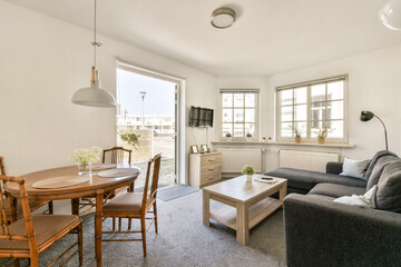 Fototapeta premium A stylish living area showcases a cozy sofa, a dining table, and large windows allowing natural light to fill the space. The design emphasizes comfort and simplicity.