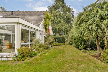 Lush greenery surrounds a modern house with large windows. The well-maintained garden features...