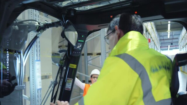 A man in a yellow jacket drives a forklift, while a woman in an orange vest and hard hat stands nearby, discussing logistics in a busy warehouse.