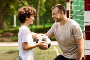 Children And Outdoor Activity Concept. Side view of smiling curly boy holding soccer ball, looking at sportive adult man who tapping and patting son on shoulder, family playing football at pitch