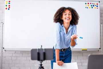 Fun English lesson and video streaming. Laughing millennial african american lady stands near whiteboard with rules and looks at camera on smartphone, talking with students online remotely, copy space
