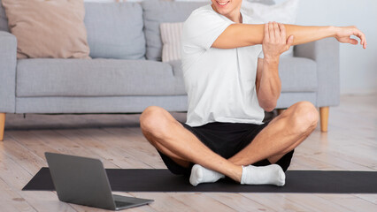 Obraz premium Online yoga lessons. Smiling young man doing warm-up and sitting in lotus pose on mat in home interior, free space
