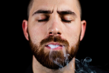 Obraz premium Pensive young caucasian male with beard exhaling smoke against black background