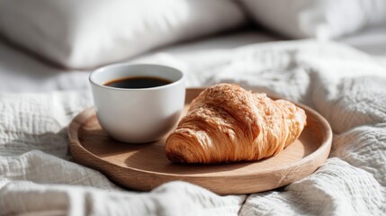 A fresh croissant and a cup of coffee are elegantly placed on a wooden tray, resting on a soft, white bedspread.