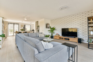 A stylish modern living room featuring a gray sofa, wooden furniture, and a wall-mounted television, illuminated by natural light from large windows.