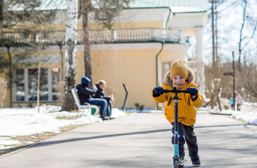 A little boy in bright yellow clothes rides a scooter. A happy child walks outside in early spring....