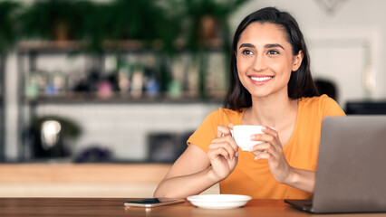 Happy young arab woman drinking coffee at cafe, using laptop, empty space