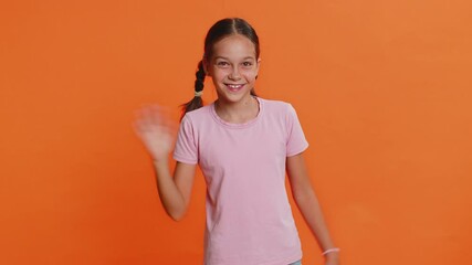 Young cute school girl 10 years old smiling friendly at camera and waving hands gesturing hello or goodbye, welcoming with hospitable expression. Preteen female child kid isolated on orange background
