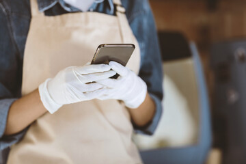 New normal, hygiene, healthcare and work during covid-19 quarantine. Young african american woman barista in apron and gloves typing on smartphone in cafe interior, cropped, close up, empty space
