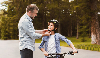 Naklejka premium Child Safety Concept. Middle age father helping his little son to put his helmet on, selective focus