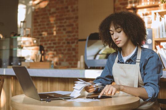 Work with accounts, social networks and online orders. Sad millennial african american lady in apron work with bookkeeping, pays bills with phone and laptop in modern cafe interior, free space