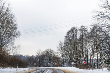 Snow-covered winter road with visible traffic signs warning of slippery conditions. The cold, icy...
