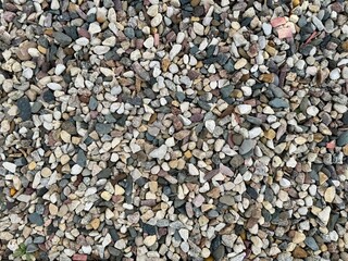  Close-up view of a diverse collection of small, multicolored pebbles scattered evenly to form a textured background, showcasing a variety of shapes and earthy tones.
