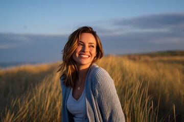 Naklejka premium A beautiful woman with a joyful smile stands amidst golden grass on a beach during the late afternoon. Sunlight enhances her features as she enjoys a serene moment by the sea
