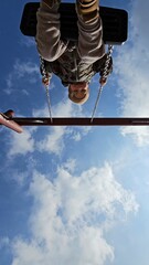 Child enjoying a swing ride captured from below, set against a backdrop of white clouds and blue sky, wearing a cosy outfit. The scene conveys a sense of freedom and happiness