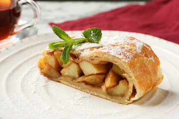 Piece of tasty apple strudel with powdered sugar and mint on table, closeup