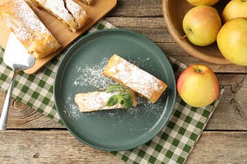 Pieces of tasty apple strudel with powdered sugar, mint, fruits and spatula on wooden table, flat lay