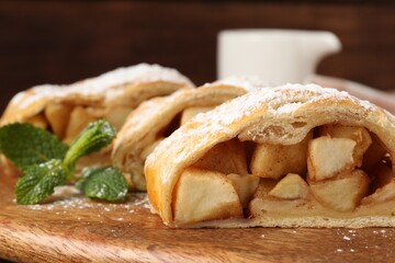 Pieces of tasty apple strudel with powdered sugar and mint on wooden board, closeup