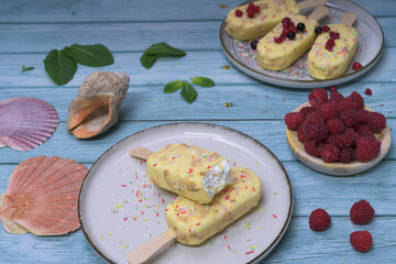 Homemade ice cream bars covered with white chocolate and red berries on a summery blue background, with raspberries and tropical decoration. Refreshing dessert concept