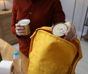 Survival kit. Woman packing different emergency supplies into backpack at table indoors, closeup