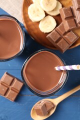 Tasty chocolate milk in glasses, pieces and banana on blue wooden table, flat lay