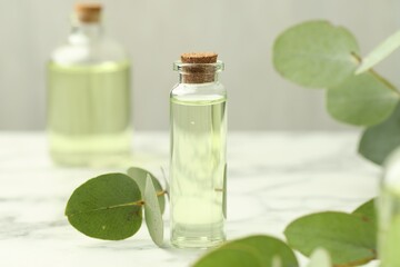 Bottle of essential oil and eucalyptus leaves on white marble table, closeup