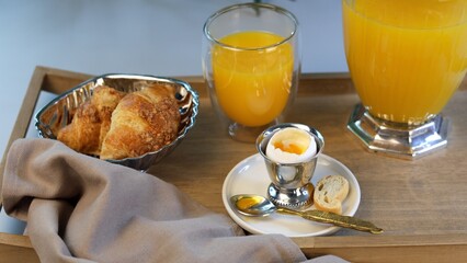 Healthy breakfast setup includes silver egg holder, golden spoon, freshly baked croissants, and vibrant orange juice, showcasing artisanal beauty in refined morning presentation.