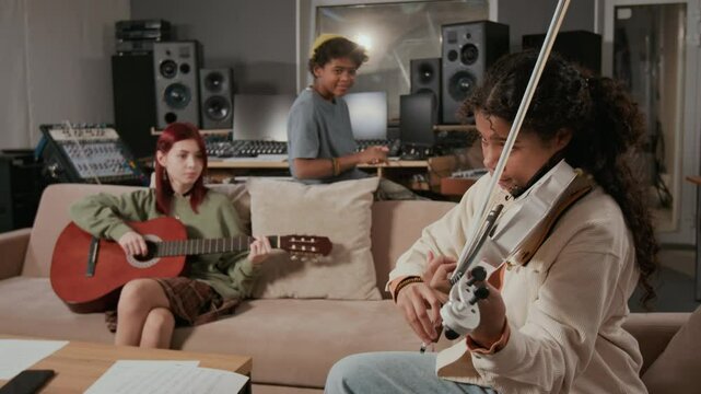 Rack focus shot of teenage girl playing violin with bow, adolescent guitarist sitting on couch, boy singer clapping along to rhythmic pattern of song in music recording studio