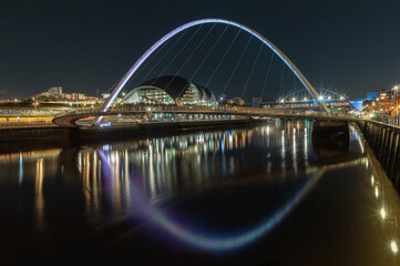 Obraz premium Night view of The Iconic gateshead millennium bridge spanning the River Tyne with Sage Gateshead with Tyne Bridge in the background. The pedestrian bridge with reflection in river, Space for text.