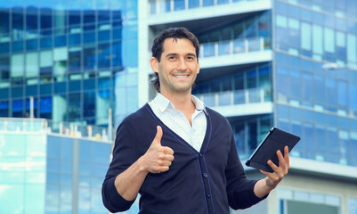 Fototapeta premium Handsome man holding pad and smiling while standing against business office building background. Thumbs up. 35 year old man.