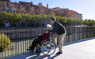 Young male caregiver spending time with an elederly woman in a wheelchair by a riverwalk on a sunny...