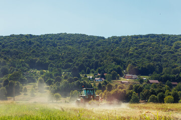 Fototapeta premium Verdant hills and dense forest tower behind a dusty field powerful tractor diligently harvests the land. Farmer cultivating and make soil tillage before seeding plants crops. Countryside rural scene