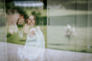 Reflection of smiling bride getting makeup done through window, surrounded by nature, preparing for...