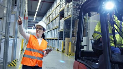 A woman in an orange safety vest directs a forklift driver in a busy warehouse, guiding the global supply chain logistics process. © wkproduction