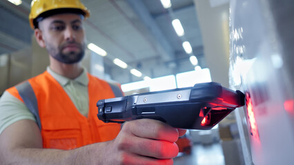 A man in a hard hat and orange vest inspects a product with a handheld scanner, ensuring quality in a warehouse setting. © wkproduction