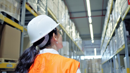 A woman in a hard hat and orange vest stands in a vast warehouse, symbolizing her role in global logistics and supply chain management. The shot emphasizes her confidence and determination.