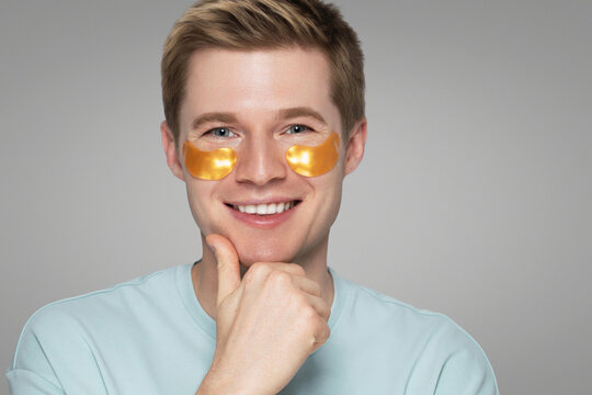 A young man with blonde hair and glowing skin smiles while wearing golden hydrogel under-eye patches and resting his hand on his chin, against a neutral grey background. Skincare for men concept.