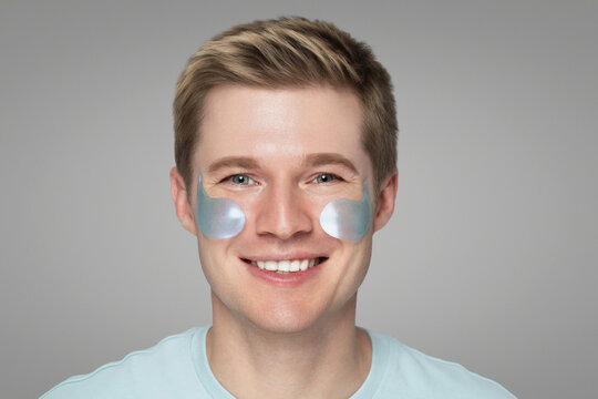 Cheerful young man with blonde hair and fresh skin smiles while wearing blue under-eye patches, standing against a neutral grey background. Men’s skincare and rejuvenation concept. - Powered by Adobe