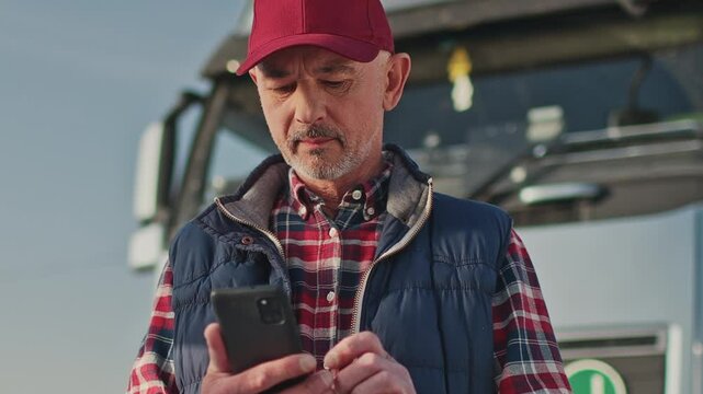 Mature Caucasian male in red cap looking down at smartphone while standing near parked truck. Concentrating on typing or reading messages. Checking routes or delivery details on screen.