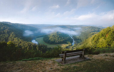 Bouillon, Belgium - Le Tombeau du Géant, panoramic view in Belgian Ardennes, Wallonia - wooden bench overlooking a scenic meander of Semois River, misty summer morning - river bent, forested hill