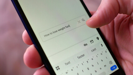 Close-up of a male hand entering the query HOW TO LOSE WEIGHT FAST into a search bar of a browser on a smartphone