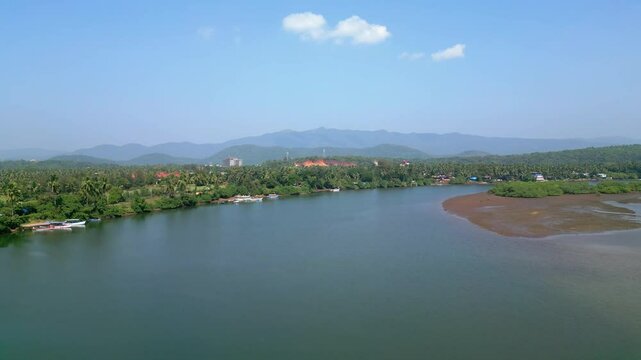 Aerial view of serene Talpona River near Rajbag Beach in Goa sparkles under the sunny sky, flanked by lush green trees and distant mountains, creating a peaceful, picturesque tropical landscape.