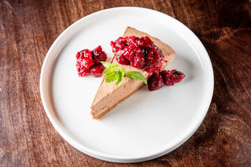 A slice of creamy chocolate cheesecake topped with vibrant raspberry compote and a fresh mint leaf. The elegant presentation enhances the dessert’s indulgent appeal.