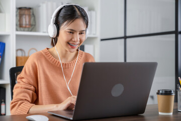 Young Asian woman students wear white headphones and use laptop to study online on the Internet at home.