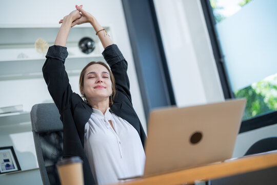 Woman stretches at her desk in a bright office space during a work break with a laptop open in front of her - Powered by Adobe