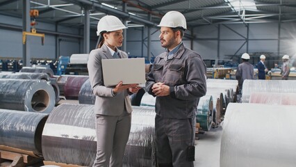 Female engineer talking with male contractor. Woman in suit and helmet holding laptop. Industry workers posing at background of metal scrolls. Manufactory employees in helmets managing work process.