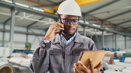 Busy African American worker accepting order from client for manufacture of industrial goods. Serious man talking on phone while entering customer data into tablet. Working with technology in plant.