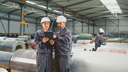 Caucasian foreman or engineer explain to his pretty assistant working process in factory. People communicating while using tablet device. In background busy workers considering industrial products.